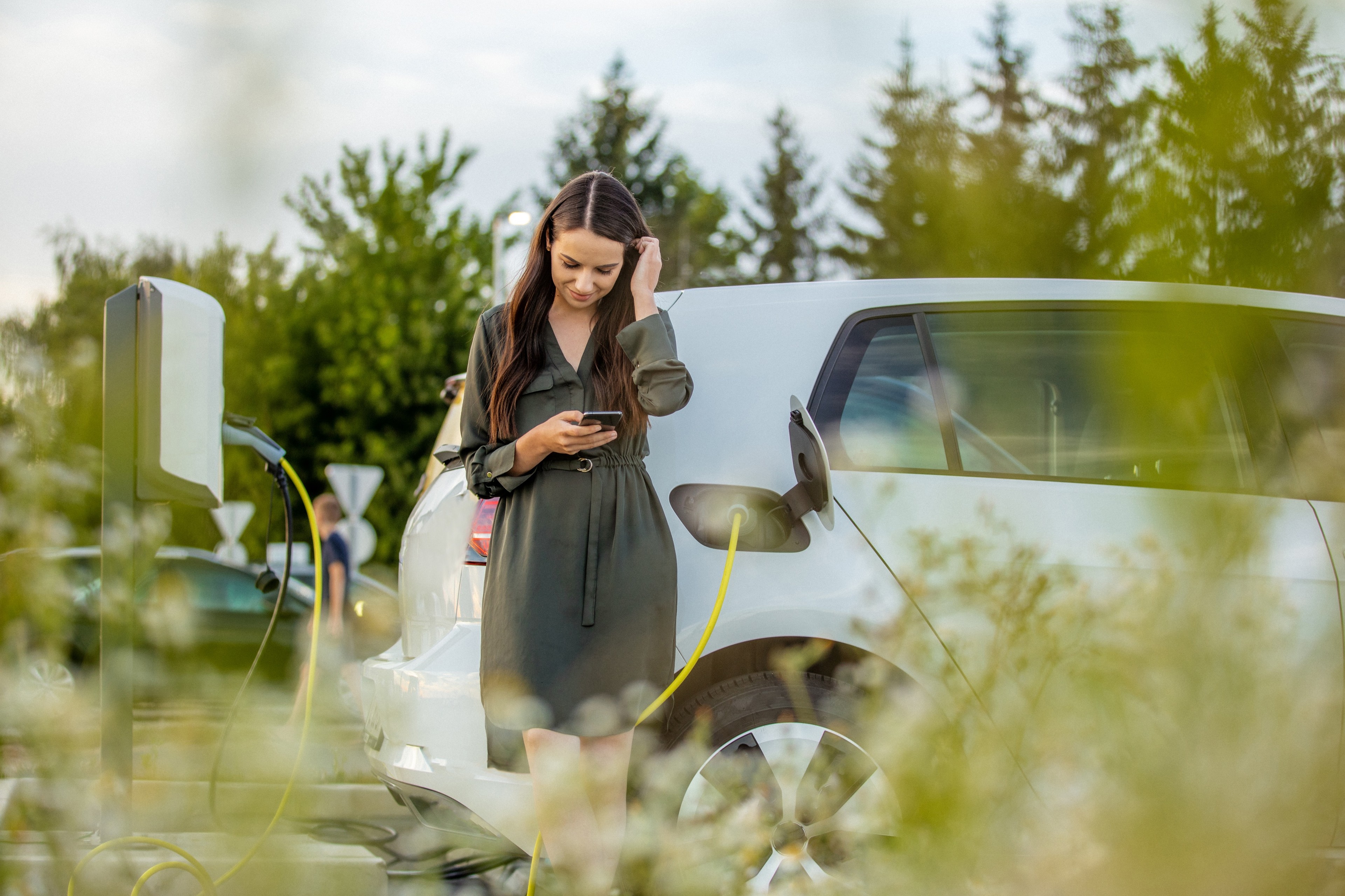 Femme rechargeant une voiture électrique tout en utilisant son téléphone.
